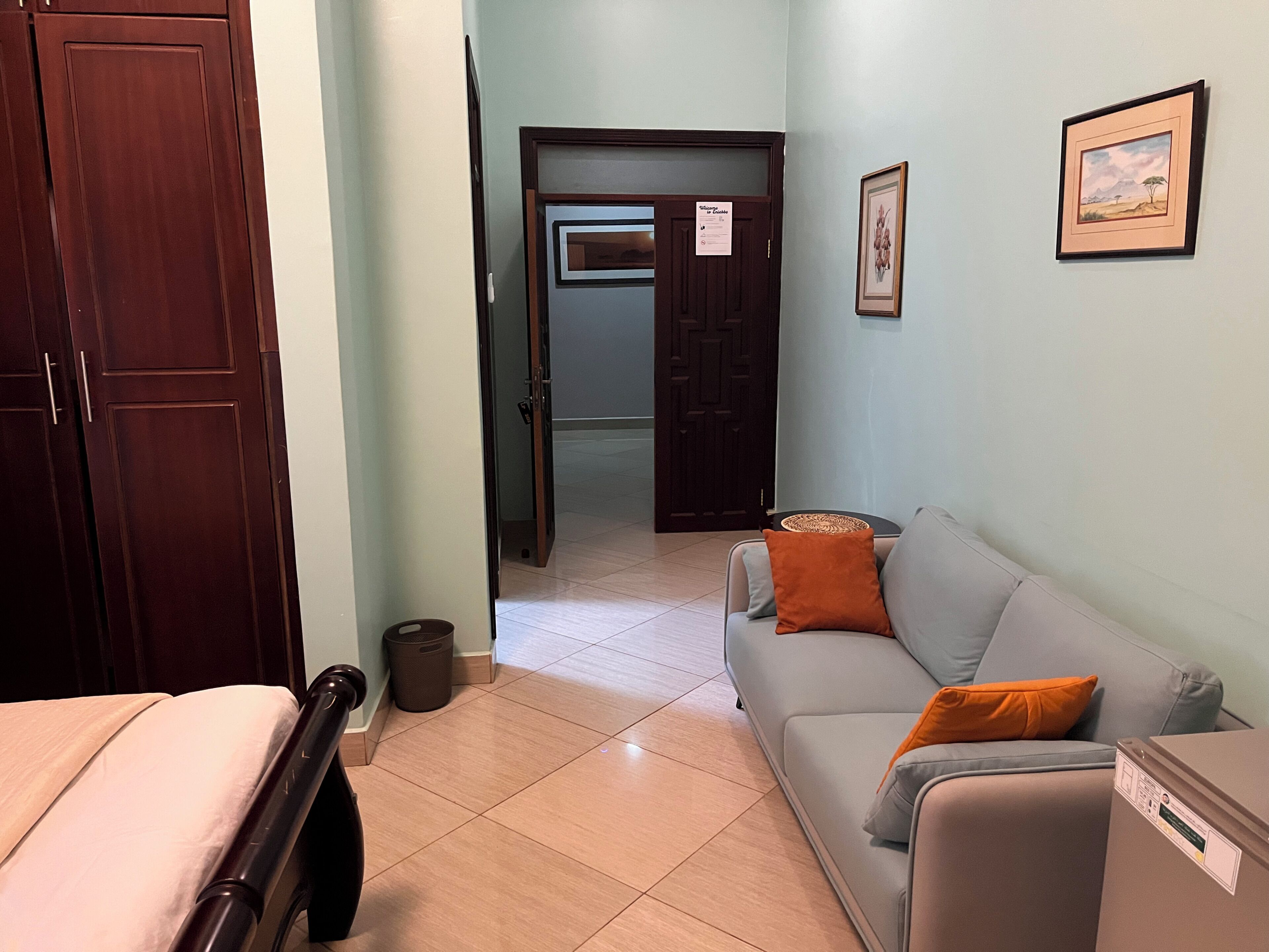 A room with a bed, a grey sofa with an orange cushion, a wooden wardrobe, and a door leading to another room.