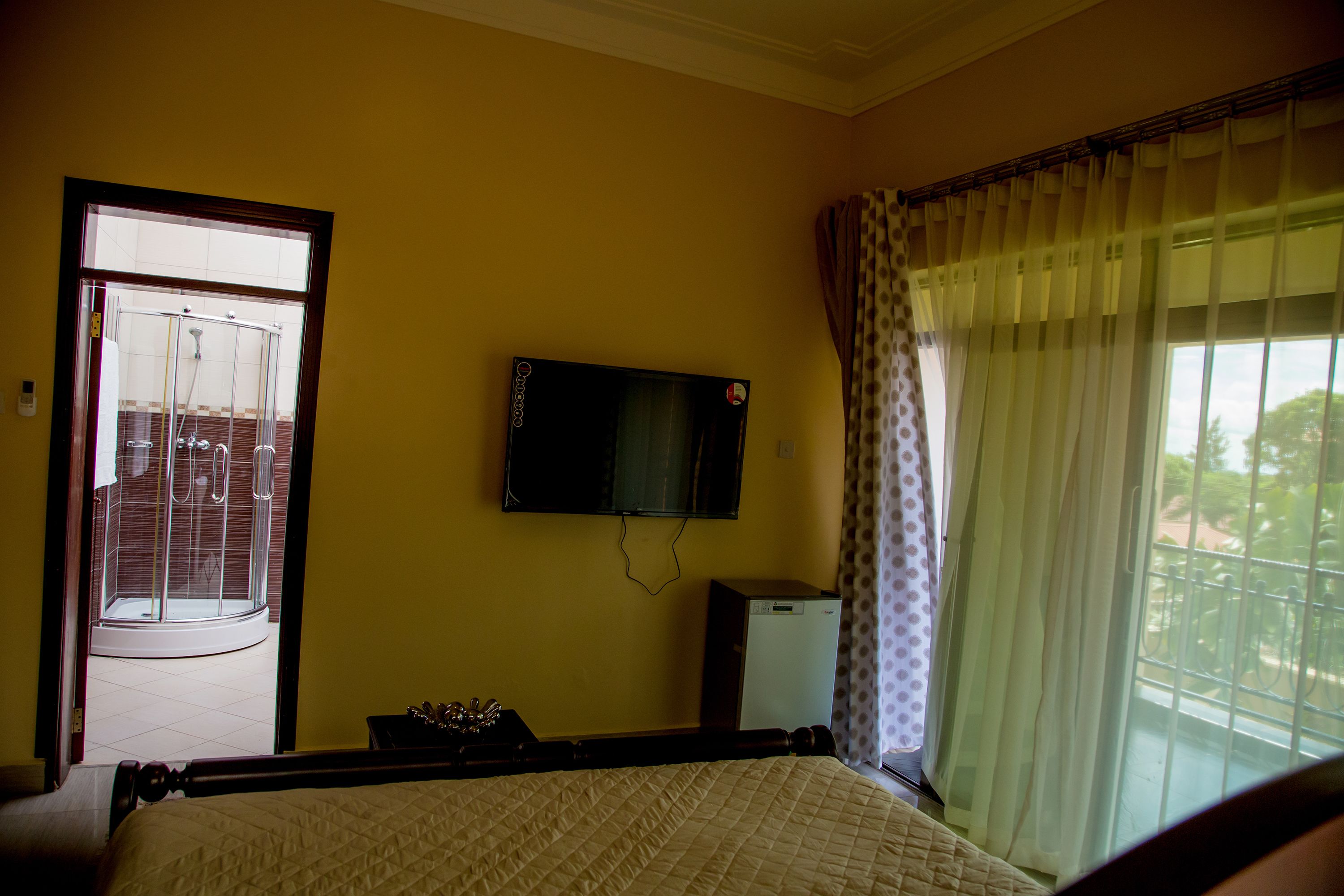 Una camera d'albergo con un letto, una televisione a muro, un piccolo frigorifero e una finestra con tende.