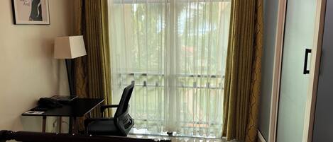 A hotel room with a bed, a desk with a chair, a lamp, and a window with curtains.