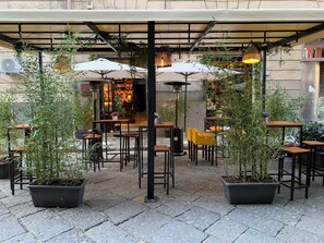 An outdoor cafe with tables and chairs, potted plants, and umbrellas.