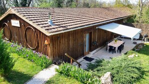 Un edificio rústico de madera con techo de tejas, un porche cubierto y un pequeño jardín con flores y arbustos.