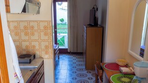 A narrow kitchen with a tiled floor, wooden cabinets, a microwave, a toaster, and a small table with a basket and a watermelon design on the tablecloth. - Eleni's Apartments & Studios (Corfu)