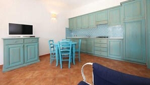 Cocina compacta con gabinetes verde mar, una mesa de comedor con sillas y un televisor sobre un mueble.