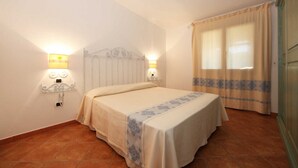 Una cama individual en una habitación con cabecera blanca, dos lámparas fijadas a la pared y una ventana con cortinas.