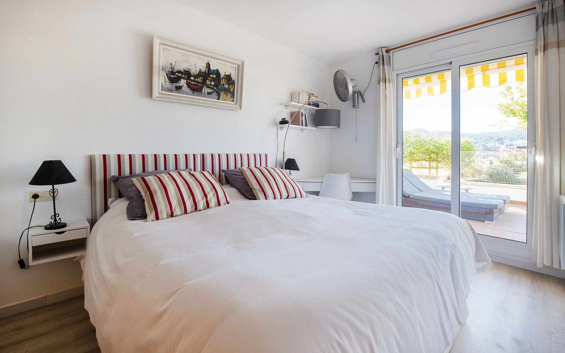 Une chambre avec un grand lit, des oreillers à rayures, une table de chevet avec une lampe, un tableau au mur et une vue sur l’extérieur par une porte coulissante.