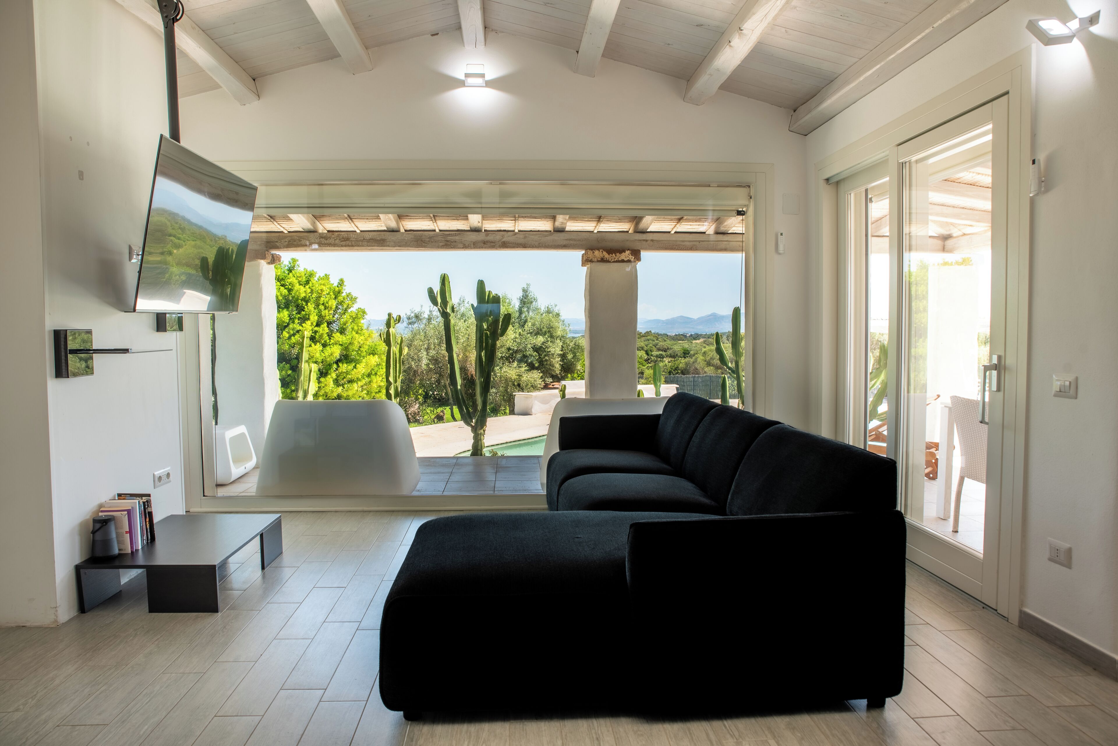 Sala de estar com sofá preto, TV de tela plana e vista para um jardim com cactos e árvores.