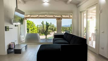 Sala de estar com sofá preto, TV de tela plana e vista para um jardim com cactos e árvores.