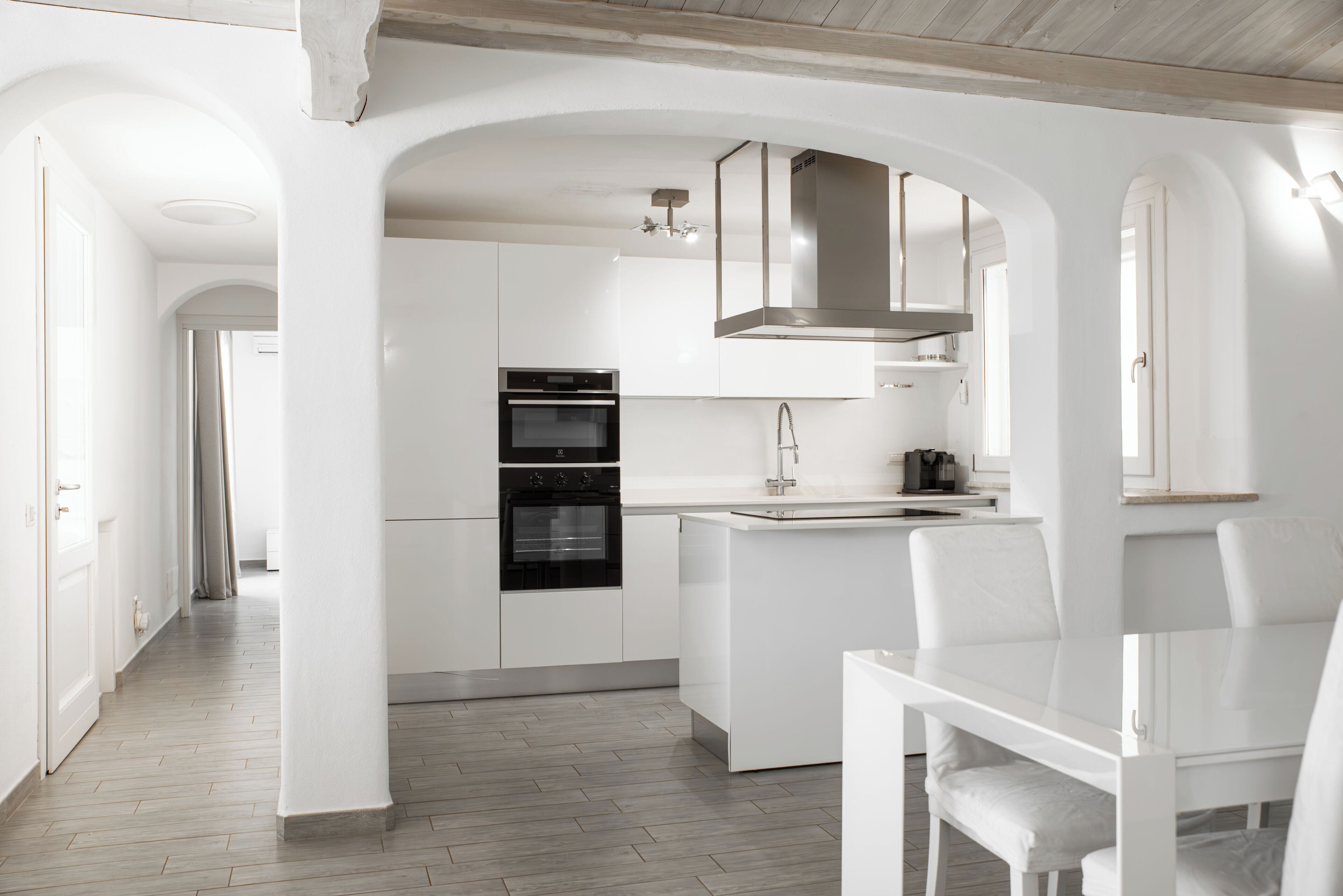 A modern kitchen with white cabinetry, a built-in oven, and a dining area with a white table and chairs.