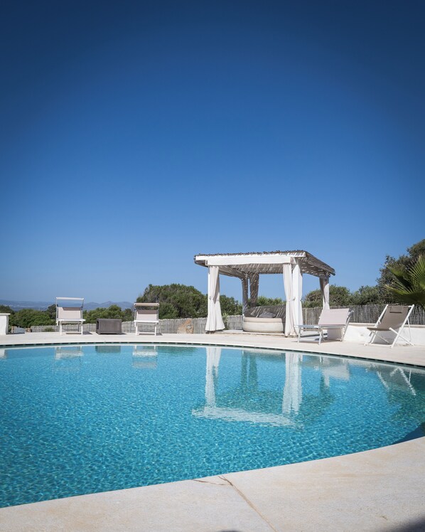 Una piscina azul cristalina con sillas de descanso blancas y una pérgola.