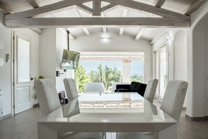 Un coin repas avec une table en verre, des chaises blanches et une vue sur des cactus et des arbres, visible à travers une porte ouverte.