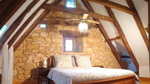 A bedroom with a stone wall, wooden beams, a bed with patterned bedding, a nightstand, a lamp, and a window. - Les Papillons 4/6, Pool, Balcony, Garden & Courtyard, Top Location & Activities (Domme)