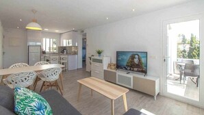 Un salon moderne avec un canapé, une table basse et un téléviseur. On y trouve également un coin repas avec une table et des chaises, et une cuisine à l’arrière-plan.