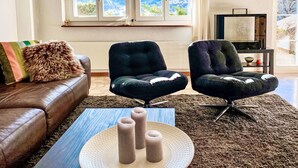 Una sala de estar con un sofá de cuero, dos sillones negros, una mesa de centro con velas y un ventanal con vistas a la montaña.