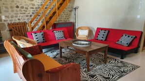 A cozy living room with a stone wall, a wooden staircase, a red sofa set with patterned cushions, a wooden coffee table, and a zebra-patterned rug. - Large stone house with enclosed garden, pets allowed (Marcillat-en-Combraille)