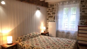 A cozy bedroom with a wooden ceiling, a quilted bedspread, a nightstand with a lamp, a dresser, and a window with a curtain. - Large stone house with enclosed garden, pets allowed (Marcillat-en-Combraille)