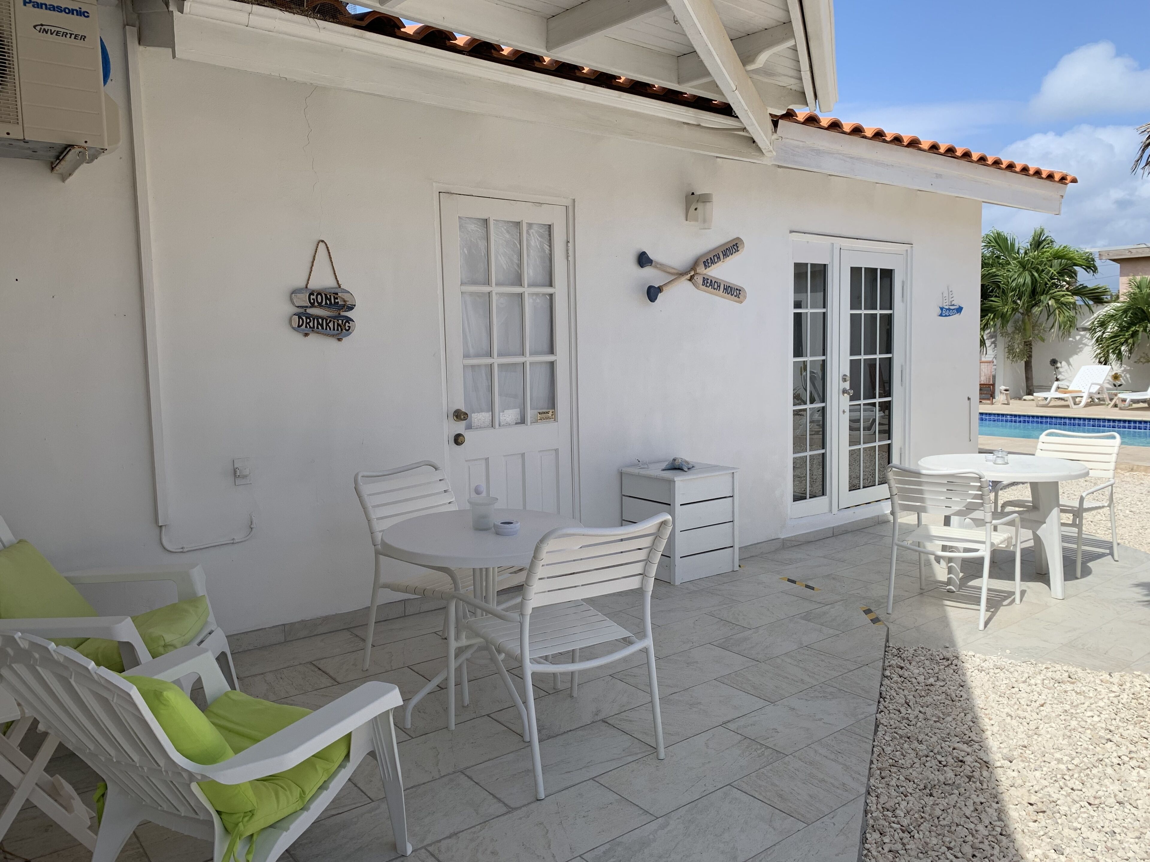 Un bâtiment blanc doté d’un patio couvert, avec des meubles blancs, une petite table et une chaise avec un coussin vert. On peut voir des panneaux sur le mur indiquant « Résidence de plage » et « On va boire un verre ».