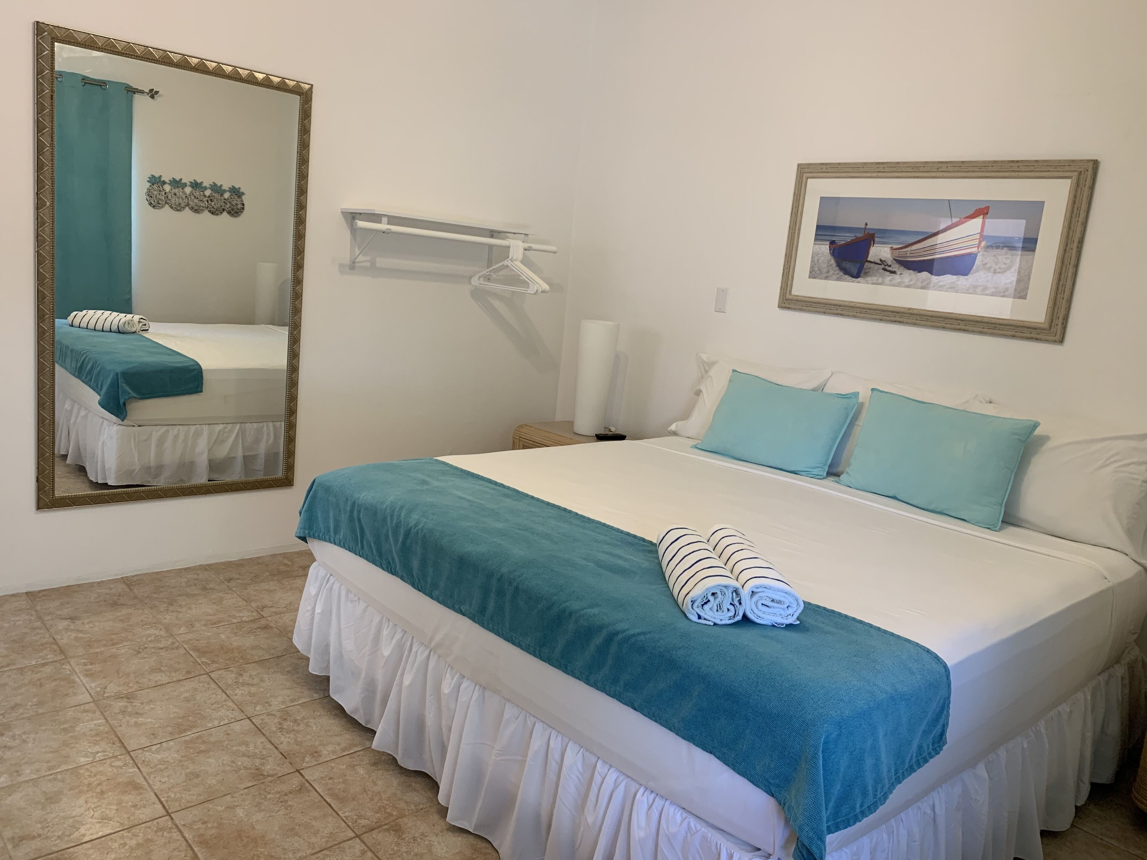 Een netjes opgemaakt bed met een blauwgroene spreidek en een witte onderrok, een grote spiegel met gouden lijst, een ingelijst schilderij van een boot aan de muur en een witte handdoek op het bed.
