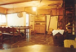 A rustic dining area with a stone floor, wooden furniture, and a kitchen in the background. - Rural house (full rental) Carriles Romanos for 8 people (Castrillo de Cabrera)