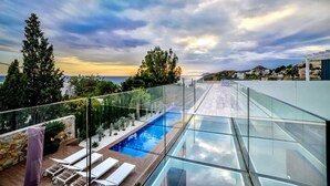 Ein moderner Pool auf dem Dach mit Glasgeländern, Liegestühlen und Blick auf das Meer und die Hügel.