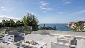 Eine Dachterrasse mit weißer Möbel, einer Glasumlaufung und Blick auf das Meer und die Hügel.