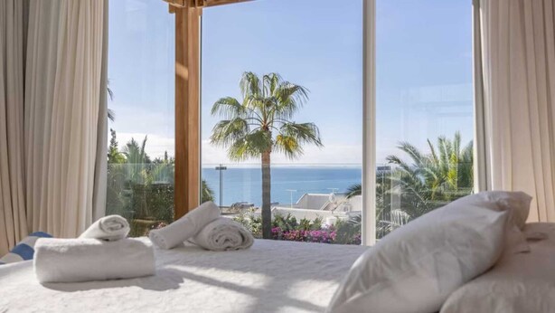 Ein Schlafzimmer mit großem Fenster, einem Bett und Blick auf das Meer und eine Palme.
