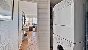 A laundry room with stacked washers and dryers, a tiled floor, and a hallway leading to a kitchen area. - Marshfront 14E Beautiful View, Beach & Pool! (Pawleys Island)