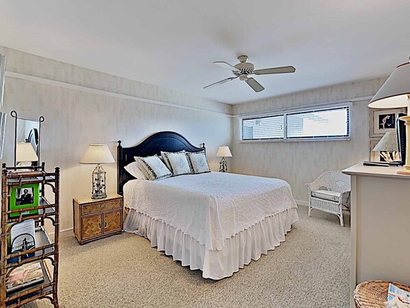 Une chambre à coucher comprenant un lit, deux tables de chevet, un ventilateur de plafond, une fenêtre avec des stores, une chaise et une petite table avec une lampe.