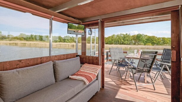 Eine Holzterrasse mit einer Couch und Stühlen, mit Blick auf ein Gewässer und Grünflächen.