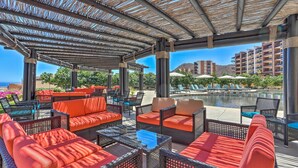 Zona relax all'aperto coperta, con mobili in vimini e cuscini arancioni, con vista su piscina e edifici dell'hotel.