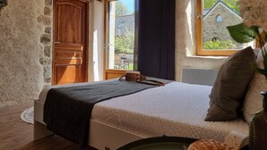 A bedroom with a stone wall, wooden door, and large windows offering a view of a stone building and greenery. - Country house by the sea - Perros Guirec on the Pink Granite Coast (Saint-Quay-Perros)