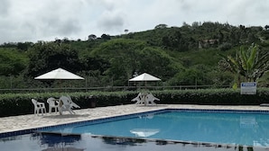 Une piscine dont le bord est revêtu de carrelage, entourée de verdure et surplombée par un ciel nuageux.