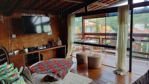 Une pièce chaleureuse avec un téléviseur fixé au mur en briques, une table en bois avec des chaises, un lit avec une literie à motifs floraux et une grande fenêtre avec des rideaux.