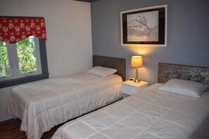 A bedroom with two beds, a nightstand with a lamp, a framed picture on the wall, and a window with a red curtain. - Charming 3-Bedroom Getaway Just 1 Mile from Sea Street Beach – Dennisport (Dennis Port)