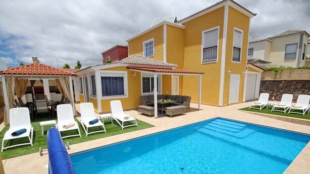 Una piscina con sillones de descanso, una zona de estar cubierta y un edificio amarillo con tejado de azulejos.