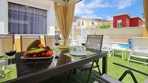 Un área de comedor al aire libre con una mesa para dos, un plato de fruta, copas de vino y vistas a una piscina y edificios al fondo.
