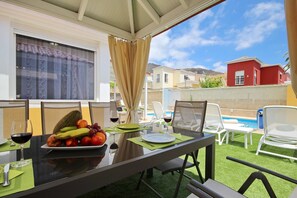 Zona pranzo all'aperto con tavolo apparecchiato per due, un piatto di frutta, bicchieri da vino e vista su piscina e edifici sullo sfondo.