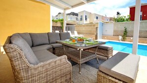Un área de estar al aire libre techada, con piscina, mobiliario de mimbre y una mesa lista para comer.