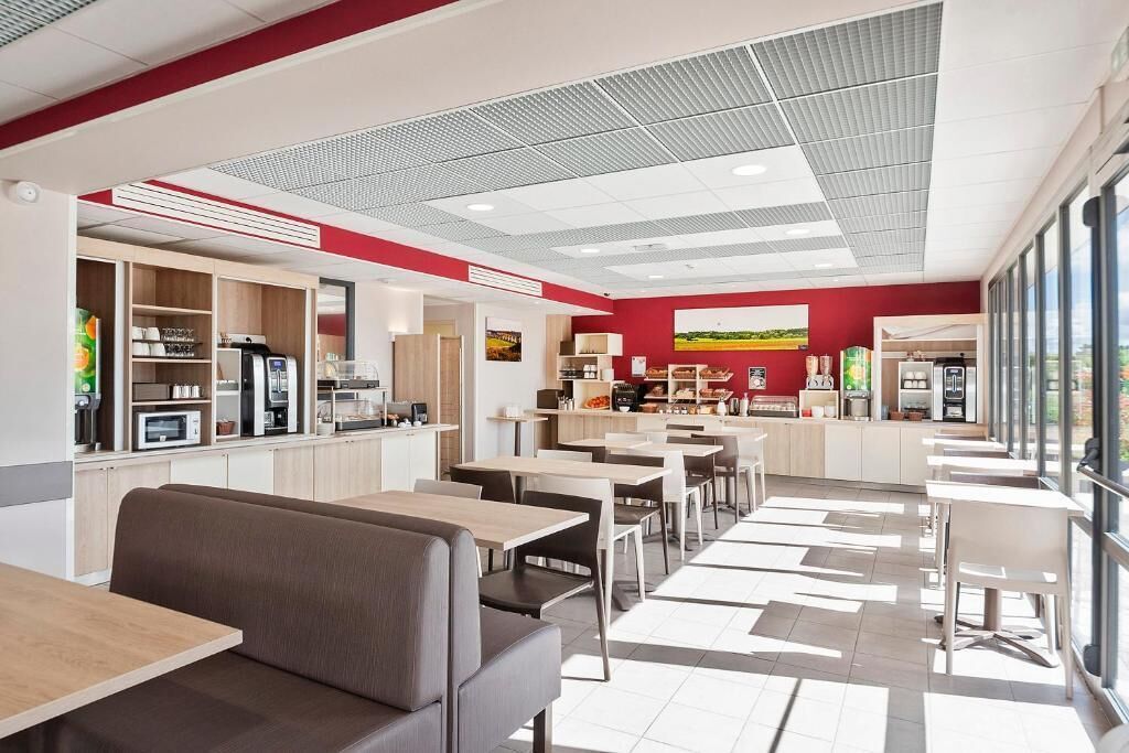 Um café moderno com áreas de estar, cozinha ao fundo e grandes janelas que permitem entrada de luz natural.