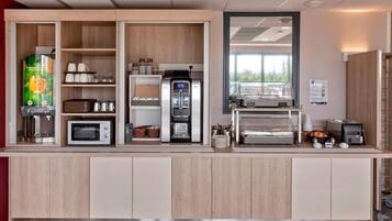 A self-service breakfast area with a coffee machine, microwave, toaster, and a display case with food items.