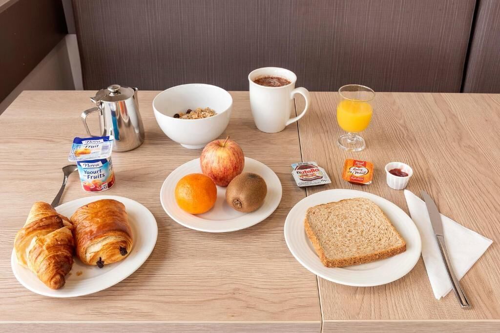 A breakfast table with croissants, fruits, a glass of orange juice, a coffee cup, and a butter packet.