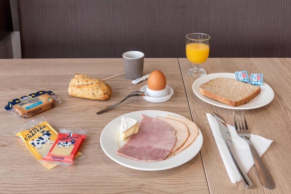 A breakfast table with a plate of ham, cheese, and bread, a glass of orange juice, a cup, and some packaged snacks.