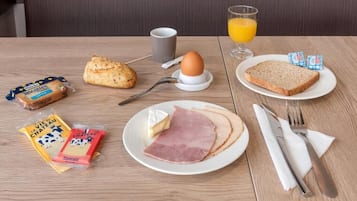 A breakfast table with a plate of ham, cheese, and bread, a glass of orange juice, a cup, and some packaged snacks.