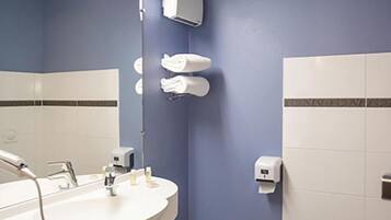 A bathroom with a white sink, mirror, and toilet. There is a trash can and a towel dispenser on the wall.
