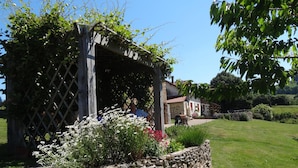 Un muro de piedra con una pérgola de madera, un edificio de piedra con techo de tejas rojas y una zona ajardinada con flores.