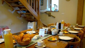 Un desayuno con una mesa de madera, sillas, una cesta de croissants, un plato de fruta, una botella de jugo y un letrero con el menú.