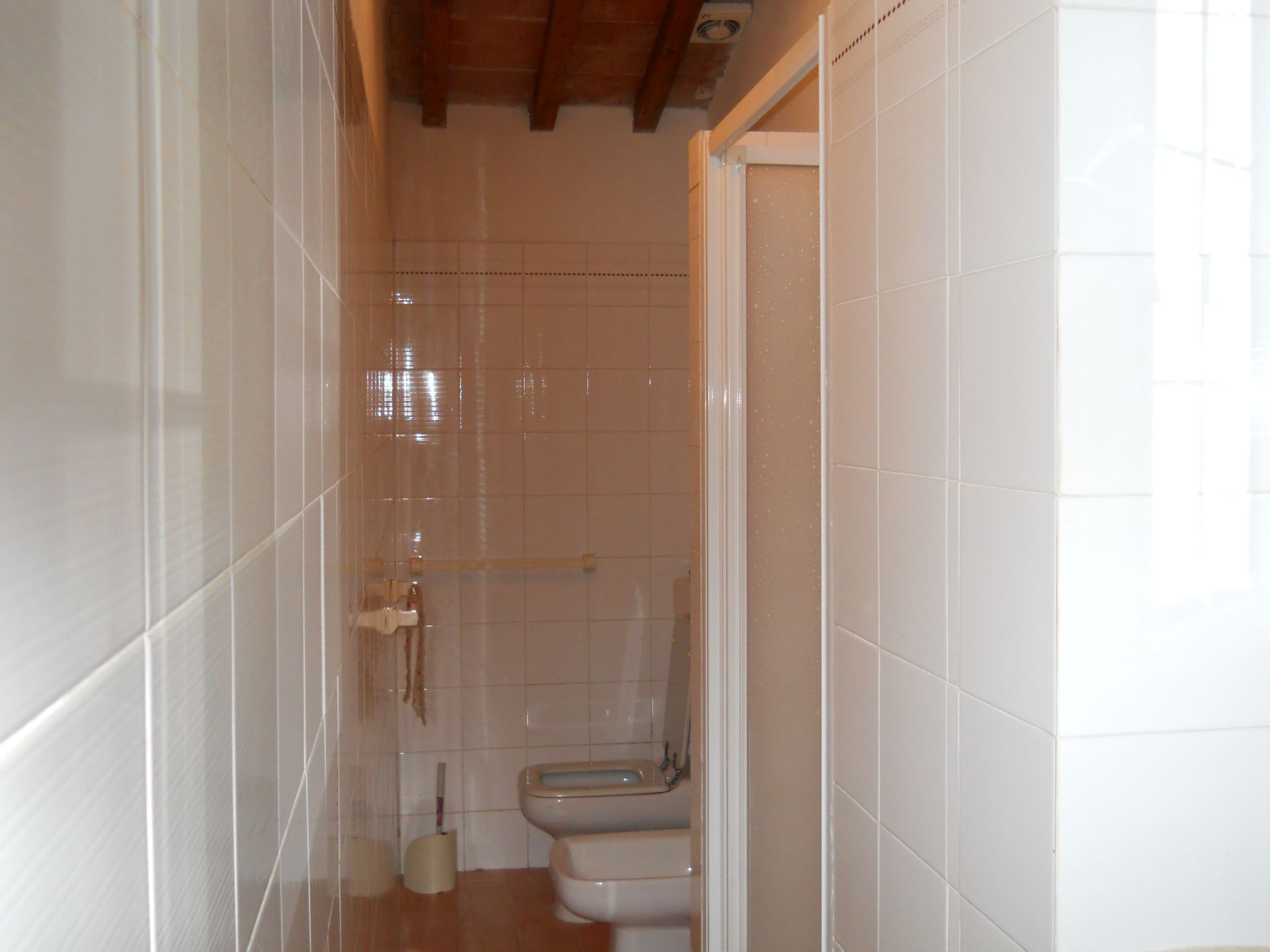 A narrow bathroom with white tiles, a toilet, and a shower area.