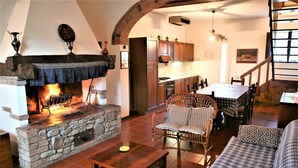 Un salon chaleureux avec une cheminée en pierre, des meubles en bois et un coin cuisine doté de placards en bois et d’une table à manger.