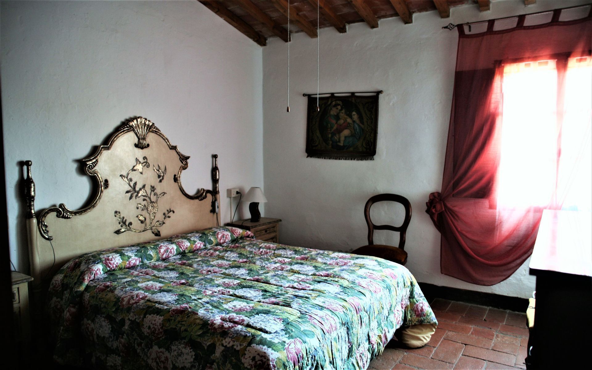 Een slaapkamer met een bed, een stoel, een nachtkastje, een lamp, een schilderij en een raam met gordijnen.