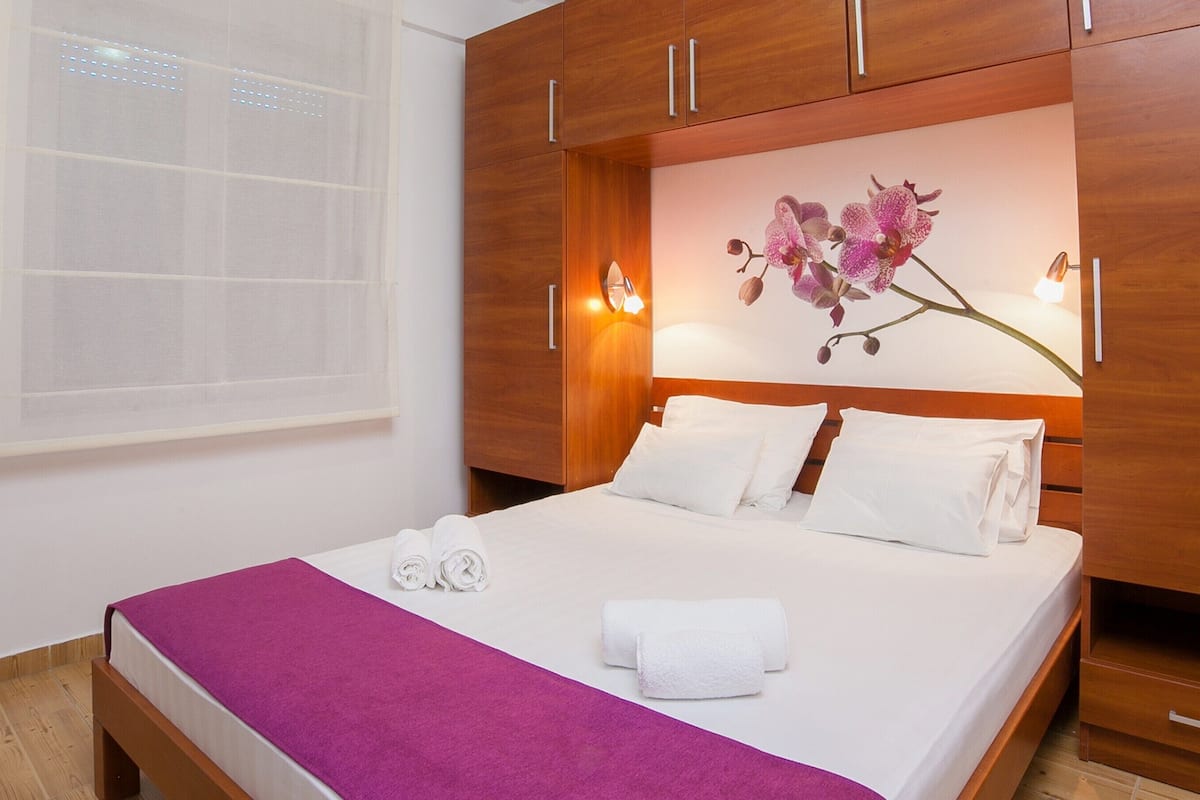 A hotel room with a bed, a purple throw blanket, white towels, and a wall-mounted orchid decoration.