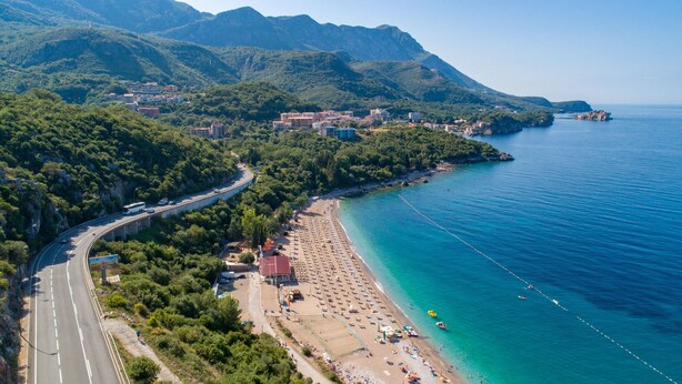 An aerial view of a coastal town with a beach, a road curving through the town, and mountains in the background. - Apartments Raymond (Pržno)